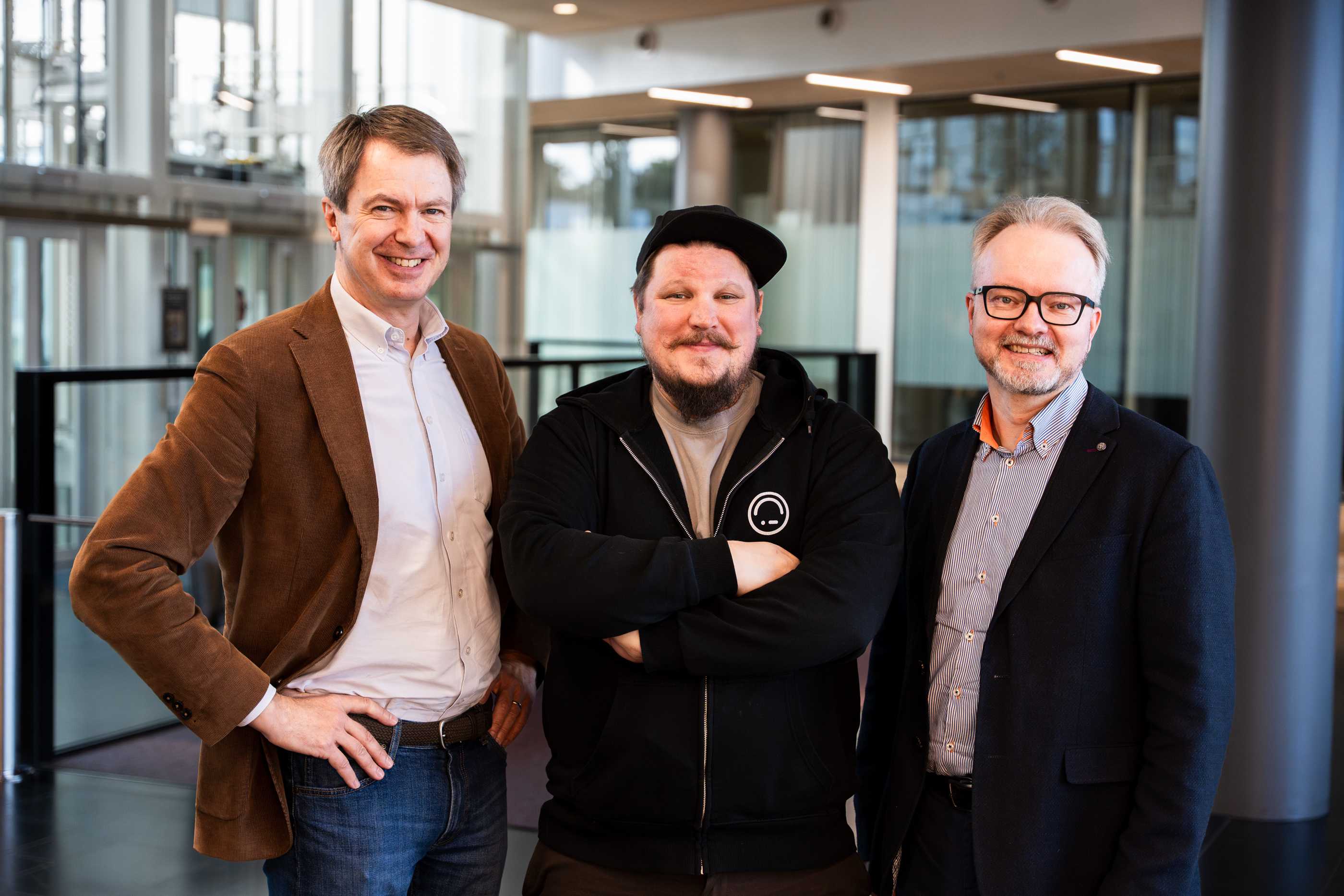 Kolme miestä seisoo yhdessä sisätiloissa hymyillen kameralle modernissa, lasiseinäisessä rakennuksessa.