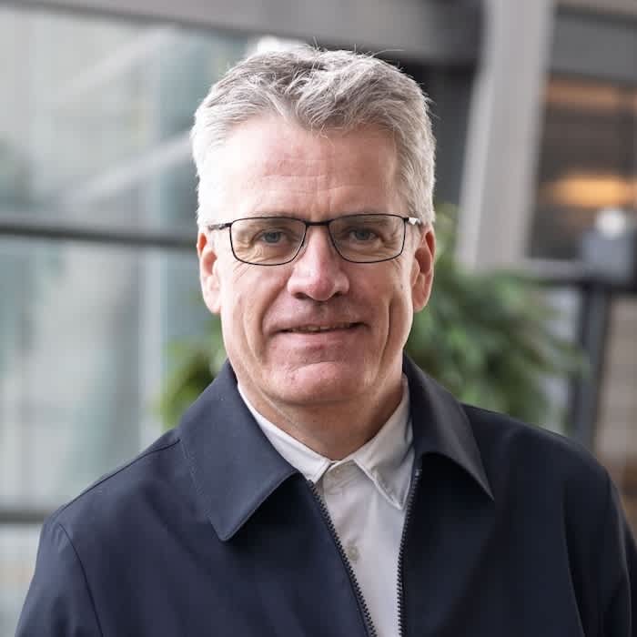 Mies, jolla on lyhyet harmaat hiukset ja silmälasit ja tumma takki, seisoo sisätiloissa, taustalla kasveja.