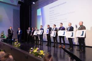 Ihmiset seisovat lavalla todistukset kädessään palkintojenjakotilaisuudessa, ja heidän takanaan on esitys.