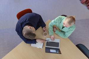 Kaksi ihmistä istuu pöydän ääressä ja työskentelee yhdessä kannettavan tietokoneen, kannettavan tietokoneen ja kahvikupin kanssa.