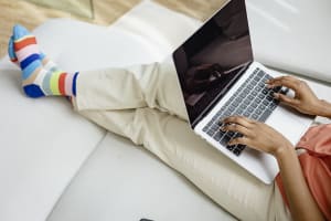 Värikkäisiin raidallisiin sukkiin pukeutunut henkilö kirjoittaa kannettavalla tietokoneella istuen vaalealla sohvalla.