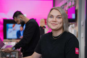 Hymyilevä mustapaitainen nainen istuu työpöydän ääressä, ja taustalla näkyy mies työskentelemässä kannettavalla tietokoneella.