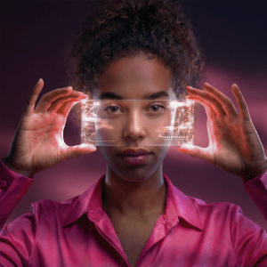 Vaaleanpunaiseen paitaan pukeutunut nainen pitää läpinäkyvää älypuhelinta kasvojensa edessä tummaa taustaa vasten.