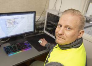 Mies keltaisissa turvaliiveissä työskentelee tietokoneella, jonka näytöllä on rakennuspiirustuksia.