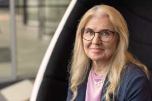 Vanhempi nainen, jolla on pitkät vaaleat hiukset ja silmälasit ja joka on pukeutunut bleiseriin, istuu ja hymyilee hieman kameralle.