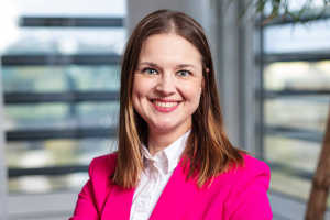 Kirkkaan vaaleanpunaiseen bleiseriin pukeutunut nainen hymyilee, kun hän seisoo sisätiloissa ja taustalla on sumeat ikkunat.