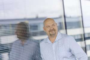 Kalju mies vaaleansinisessä paidassa seisoo kaihtimella varustetun ikkunan ääressä, hänen heijastuksensa näkyy lasissa.