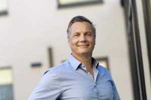 Hymyilevä vaaleansiniseen paitaan pukeutunut mies seisoo ulkona modernin rakennuksen edessä ja katsoo kameraan.