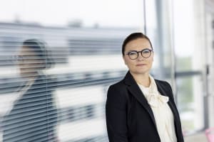 Bisnesasuun pukeutunut nainen seisoo kaihtimien varustetun ikkunan luona, hänen heijastuksensa näkyy lasissa.