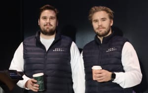 Kaksi miestä, joilla on samanlaiset liivit ja kahvikupit kädessään, seisovat sisätiloissa kameraan päin.