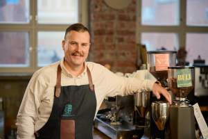 Esiliinaan pukeutunut mies hymyilee kahvimyllyjen vieressä viihtyisässä kahvilassa, jossa on tiiliseinät ja suuret ikkunat.