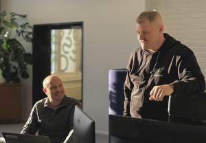 Kaksi miestä toimistossa, toinen istuu ja hymyilee toiselle, joka seisoo ja puhuu, molemmat tietokoneiden ääressä.