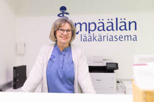 Hymyilevä silmälasipäinen nainen seisoo lääkärin vastaanotossa, jonka takana on suomenkielinen kyltti.