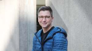 Silmälasipäinen ja siniseen takkiin pukeutunut mies hymyilee seisoessaan vaaleaa betoniseinää vasten.