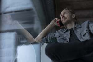 Parrakas mies puhuu punaiseen puhelimeen istuessaan sisätiloissa heijastavan lasi-ikkunan äärellä.