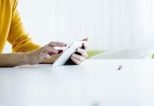 Keltaiseen paitaan pukeutunut henkilö käyttää tablettia valkoisen työpöydän ääressä valoisassa, modernissa ympäristössä.