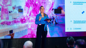 Siniseen puseroon pukeutunut nainen puhuu lavalla tabletin kanssa, etualalla suuri värikäs näyttö ja yleisö.