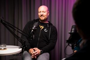 Kyberasiantuntija Antti Kurittu istuu podcast-studiossa musta huppari päällä ja hymyilee kameralle.