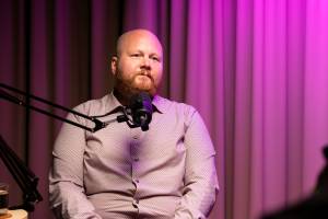 Kalju, parrakas mies istuu podcast-mikrofonin ääressä, ja hänen takanaan on violetti valaistus ja verhot.