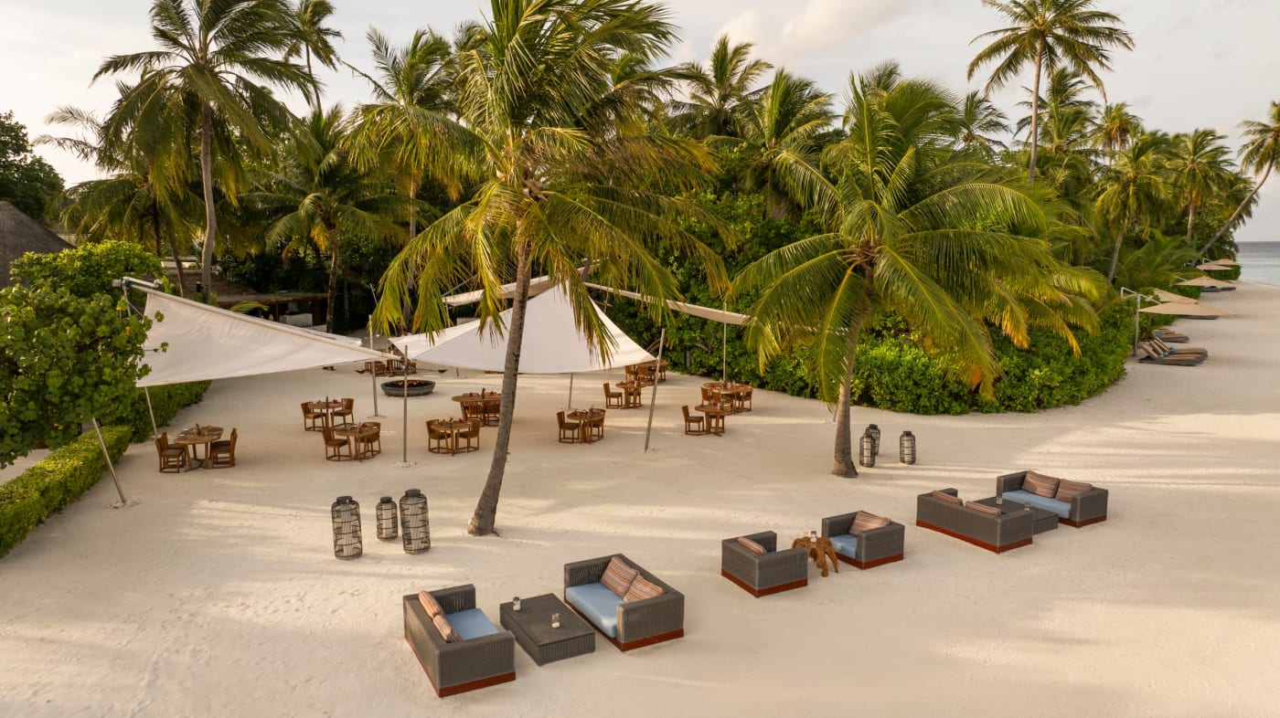Mesas y zonas de descanso dispuestas sobre la arena bajo las palmeras en el Constance Halaveli Maldives