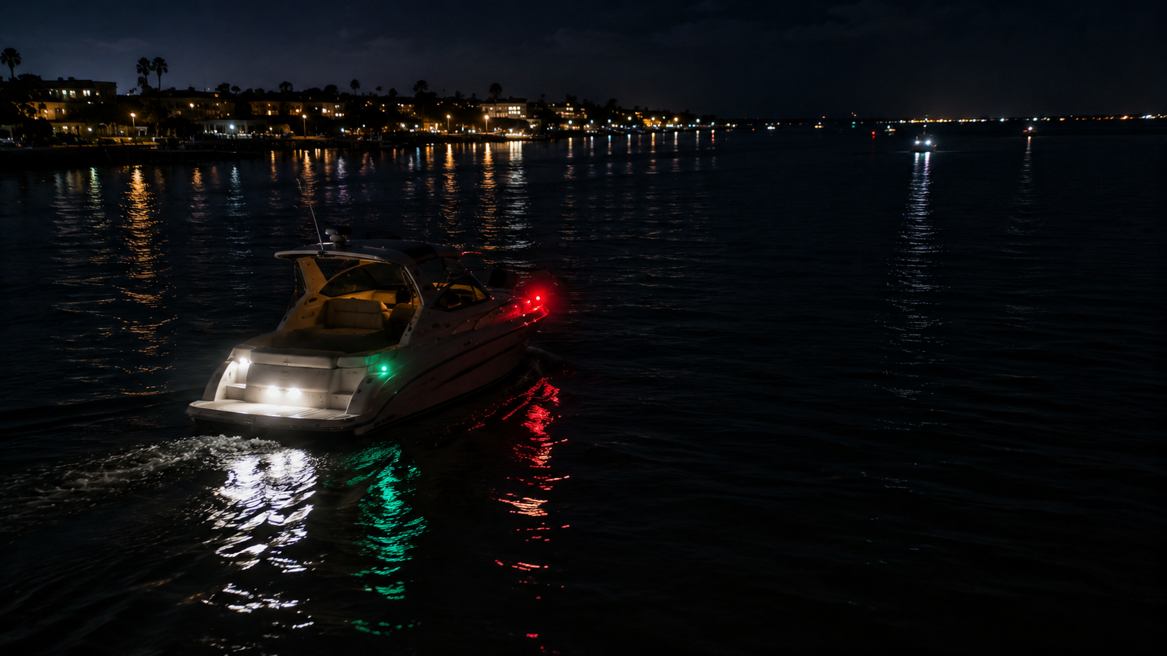 Powerboat showing red and green navigation lights crossing dark water toward a lit shoreline at night