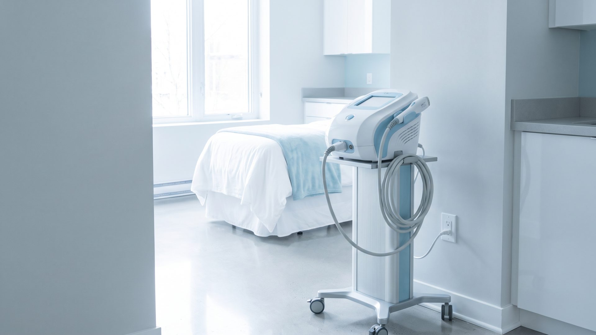 A treatment device standing on its trolley with the power cable coiled and the wall socket beside it empty, in a made-up treatment room