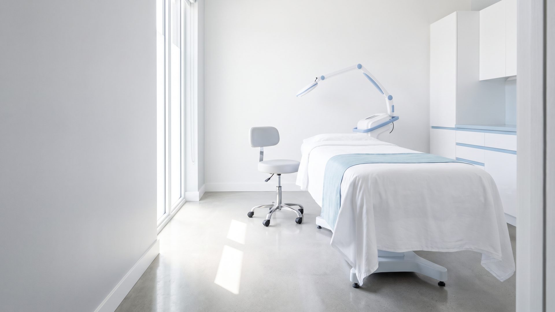 A newly finished treatment room in a medical aesthetics clinic, bed made up in white linen, morning light across the floor