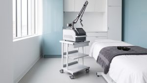 A tattoo-removal laser on its trolley beside a treatment bed, dark protective goggles resting on a charcoal towel