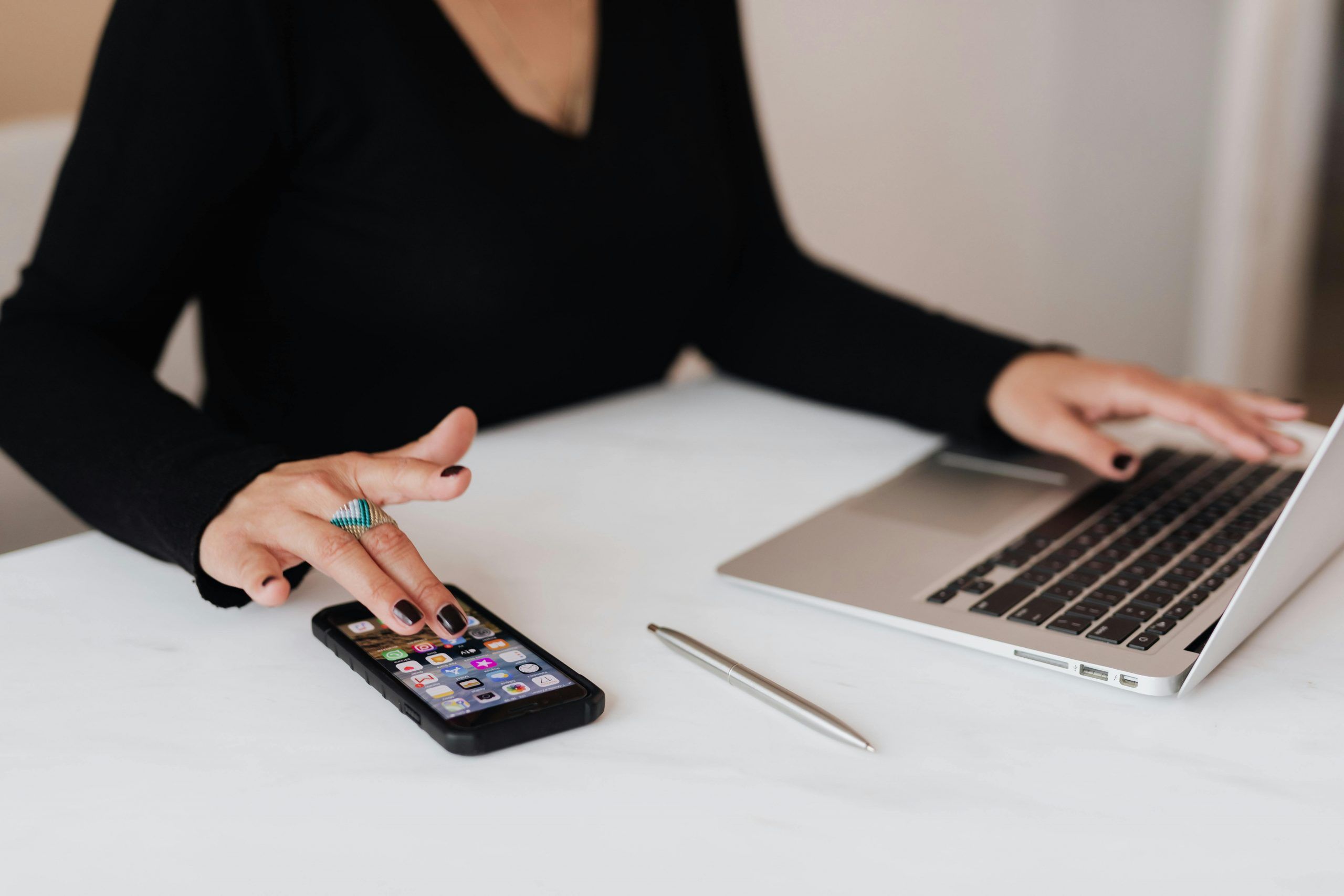 A person in a black top using a smartphone with one hand while the other rests on a laptop, with a silver pen placed on a white table.