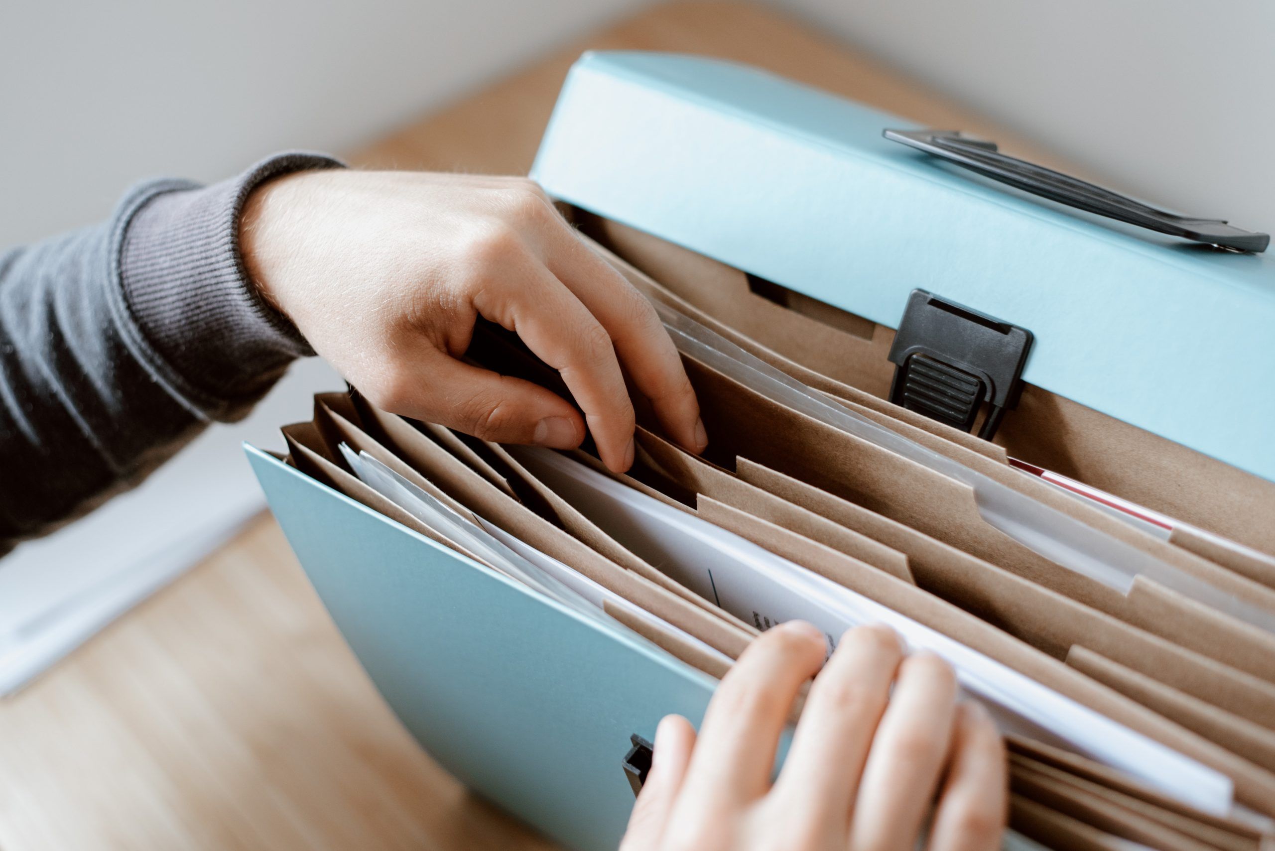 Hands searching through a blue accordion file organizer filled with papers and folders, placed on a wooden surface.