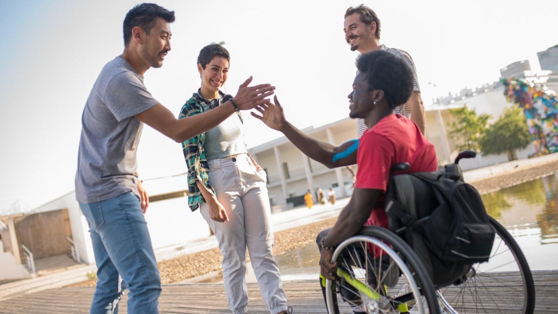 A group of friends outdoors, smiling and interacting, with one person in a wheelchair reaching out for a handshake with another
