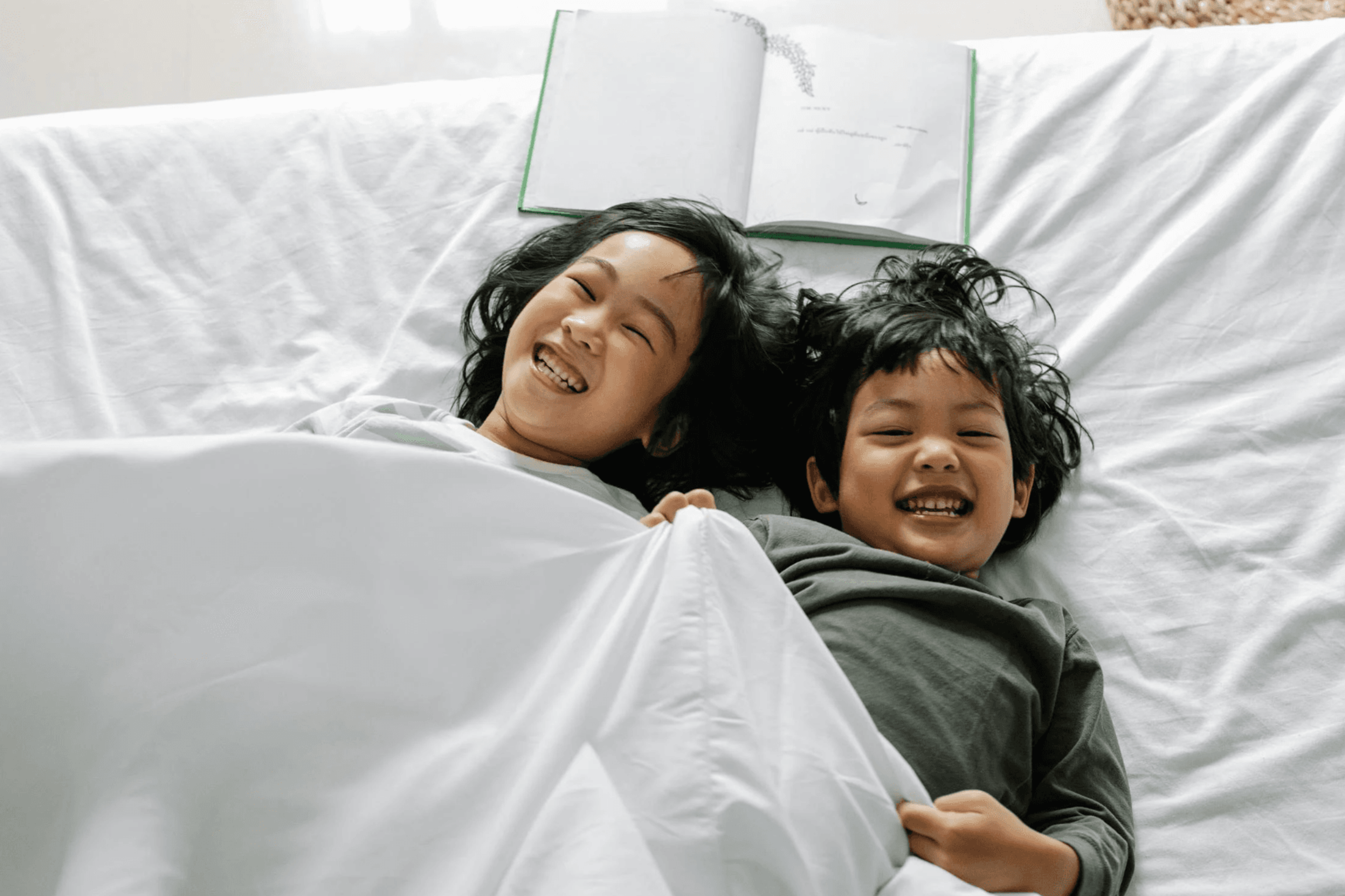 Two siblings lying on a bed, smiling and laughing under a white blanket, with an open book placed above them on the bed.