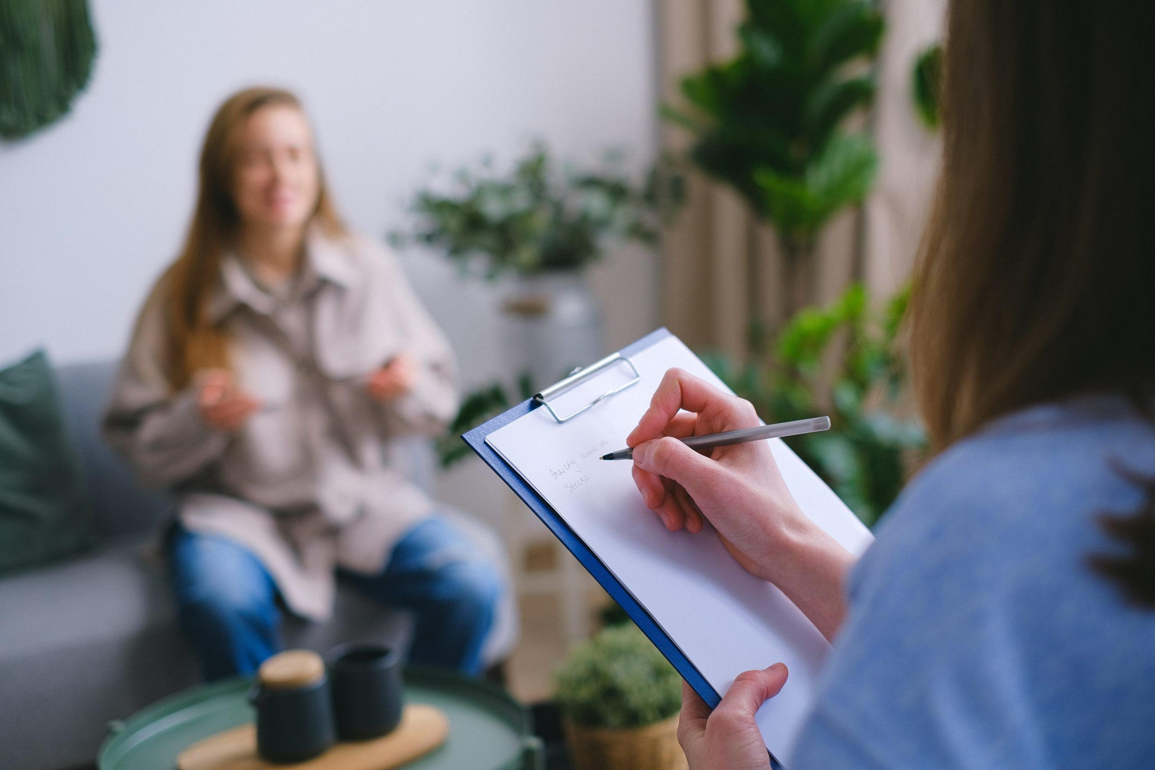 A support coordinator sitting across from a participant, holding a clipboard and pen, taking notes during a collaborative discussion in a comfortable, plant-filled room.
