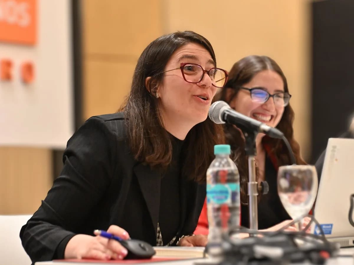 Paula Bria con micrófono en la mesa, moderando un panel junto a otra oradora