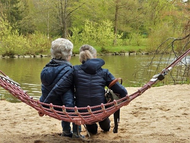 two older women sitting on a hammock in front of a pond