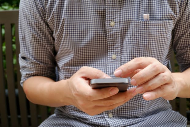 A guy in a checkered shirt is using a cell phone to look up Cialis resources while sitting on a bench outside.