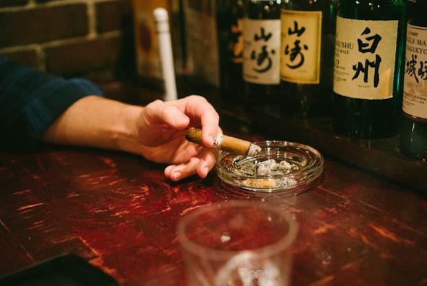 A hand holding a cigar above an ashtray in front of alcohol bottles