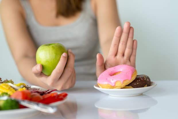 a woman holding an apple in one hand and rejecting a plate of donuts with the other hand.
