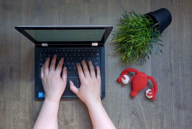 a woman typing on a computer next to a uterus made of yarn