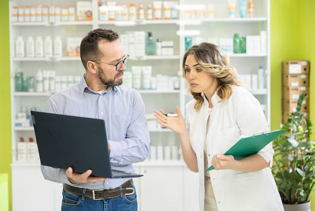 a man holding his laptop talking to a woman holding her clipboard at a pharmacy