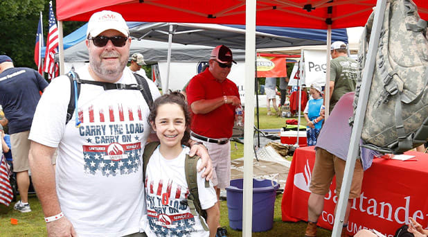 A man is standing with a child at a veterans event, both are wearing a Santander carry the load shirt.
