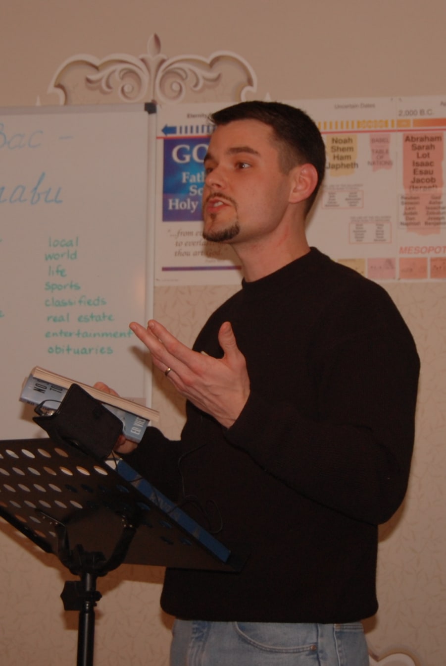 Joshua teaching from a book at a music-stand lectern in front of a Bible timeline chart
