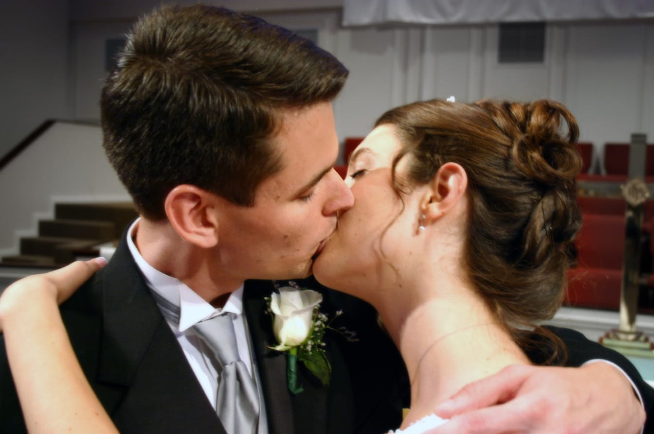 Joshua and Kelsie kiss at their wedding, the groom wearing a white rose boutonniere