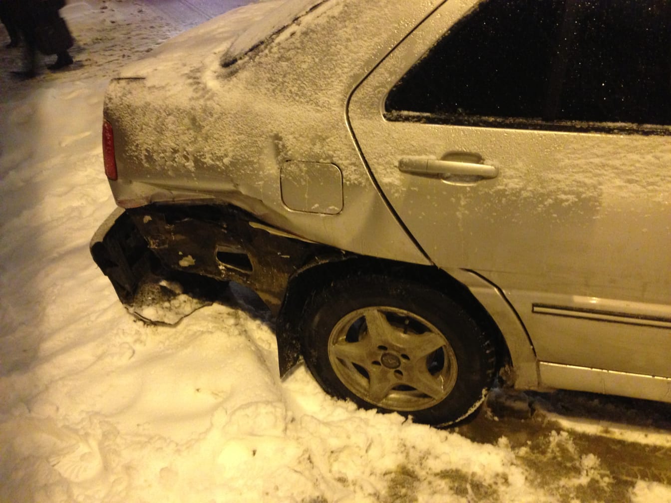 Silver car at night in the snow with its rear bumper torn away after the collision
