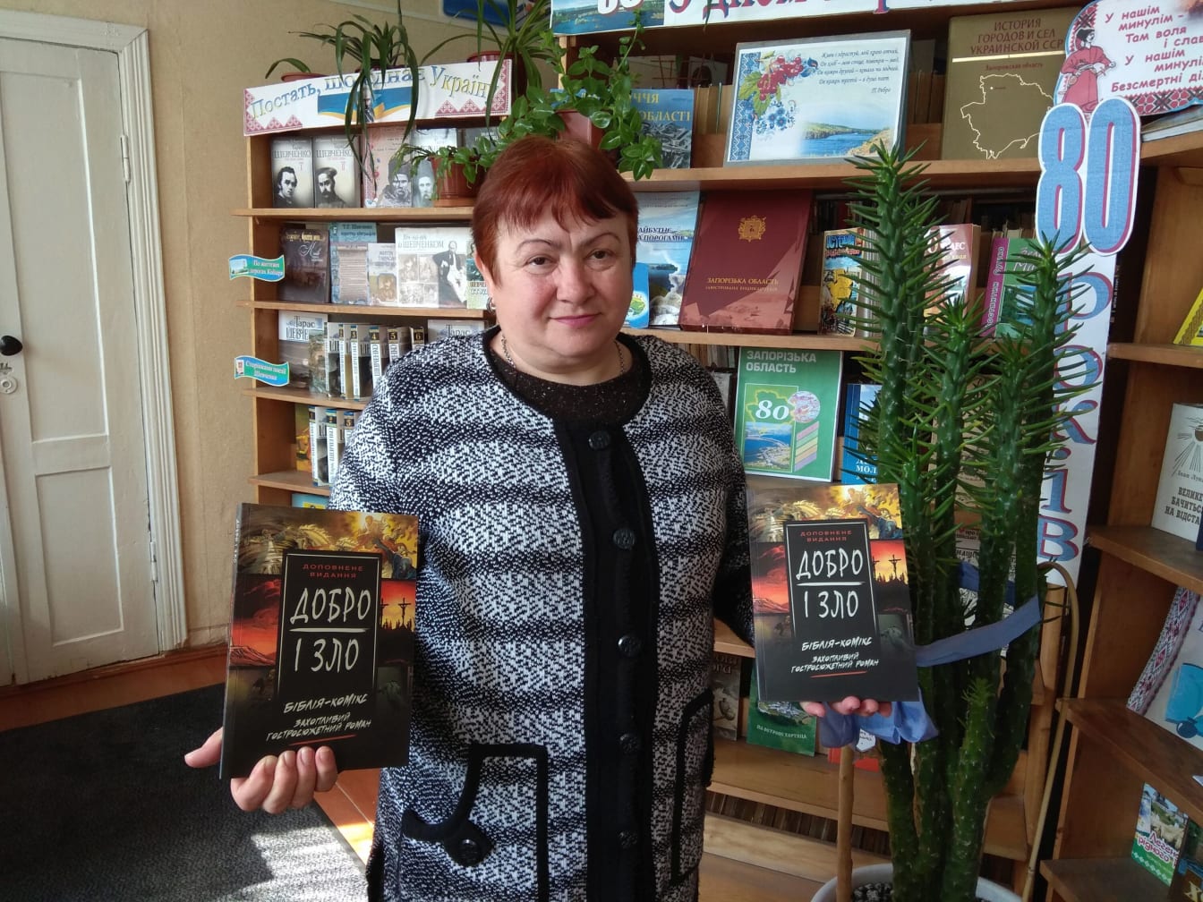 A woman holds a copy of Good and Evil, with another copy displayed on the library shelf behind her