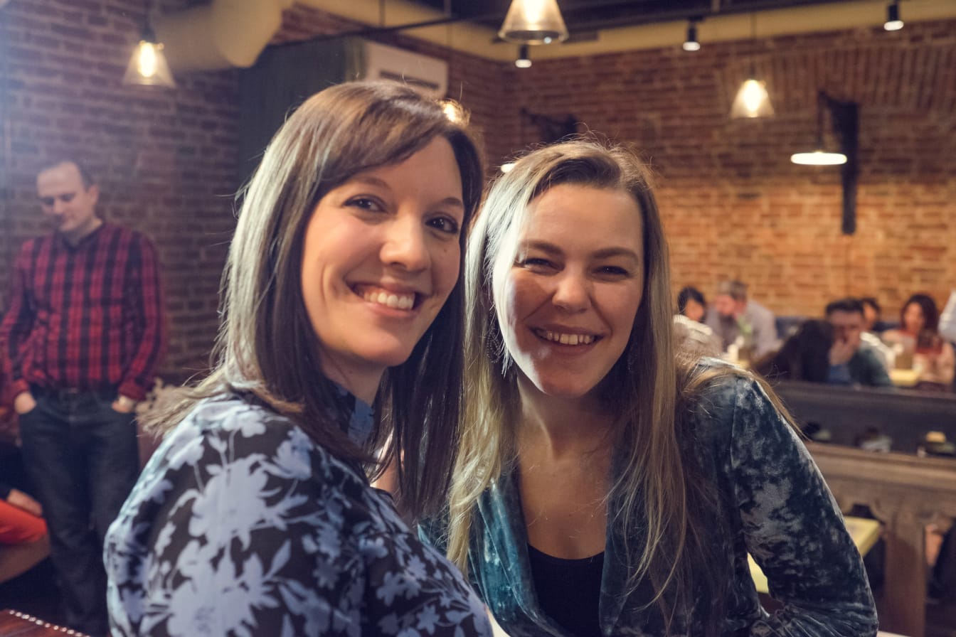Two women smiling cheek to cheek with the busy cafe behind them