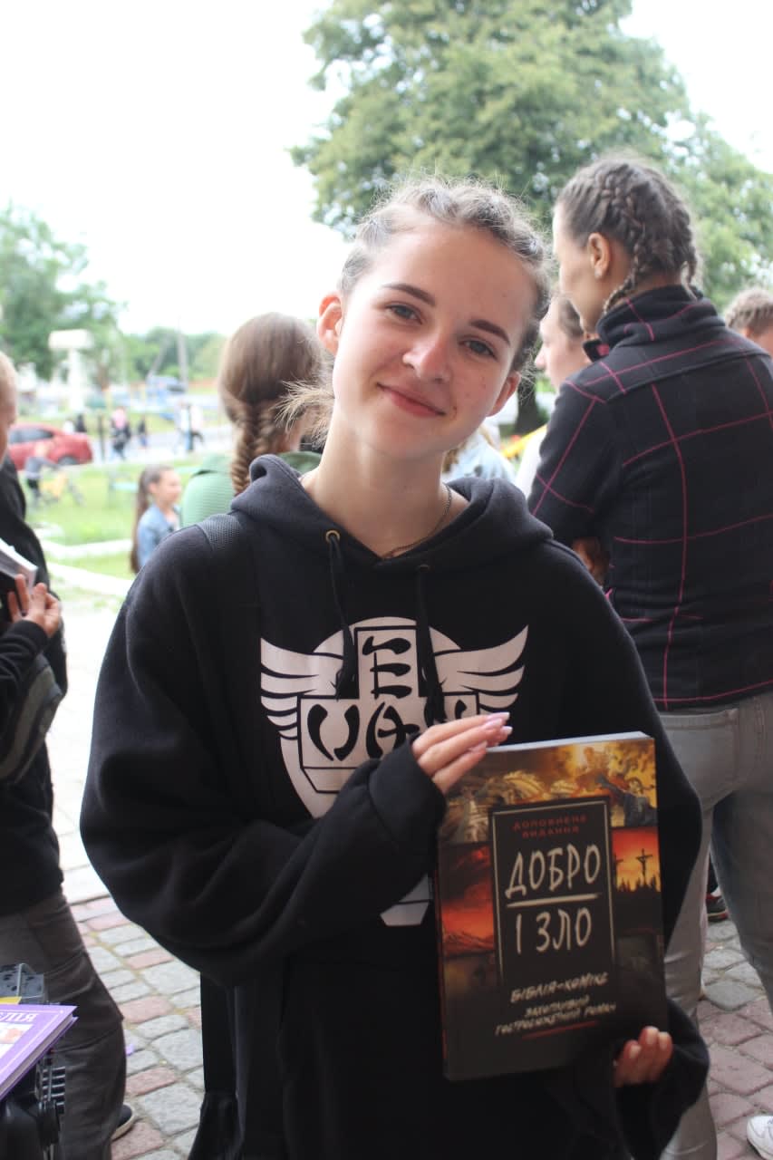 A smiling teenage girl outdoors holds a Добро і Зло (Good and Evil) book in her hands