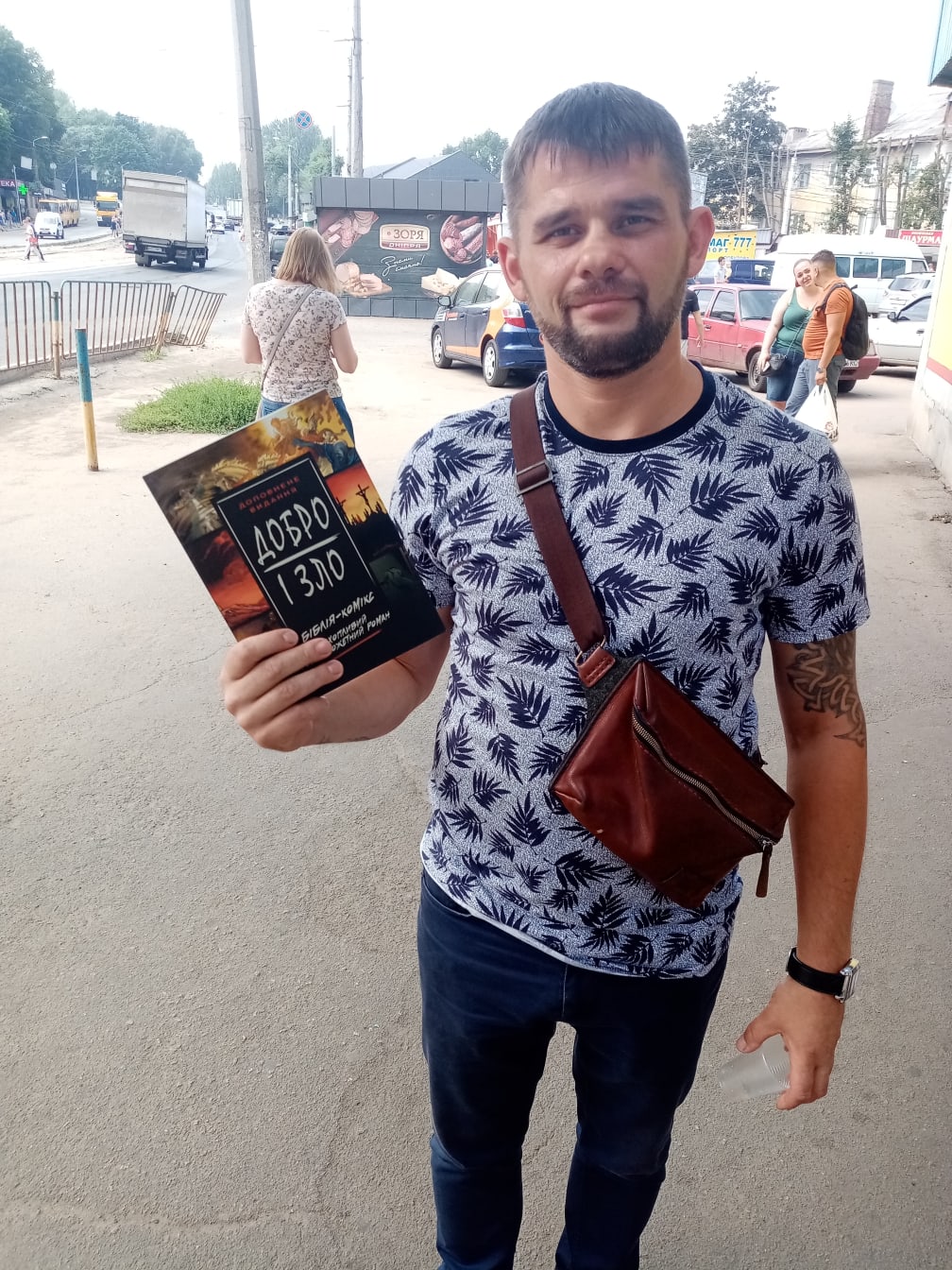 A bearded man on a city street holds up a Добро і Зло (Good and Evil) book with its dark cover facing the camera