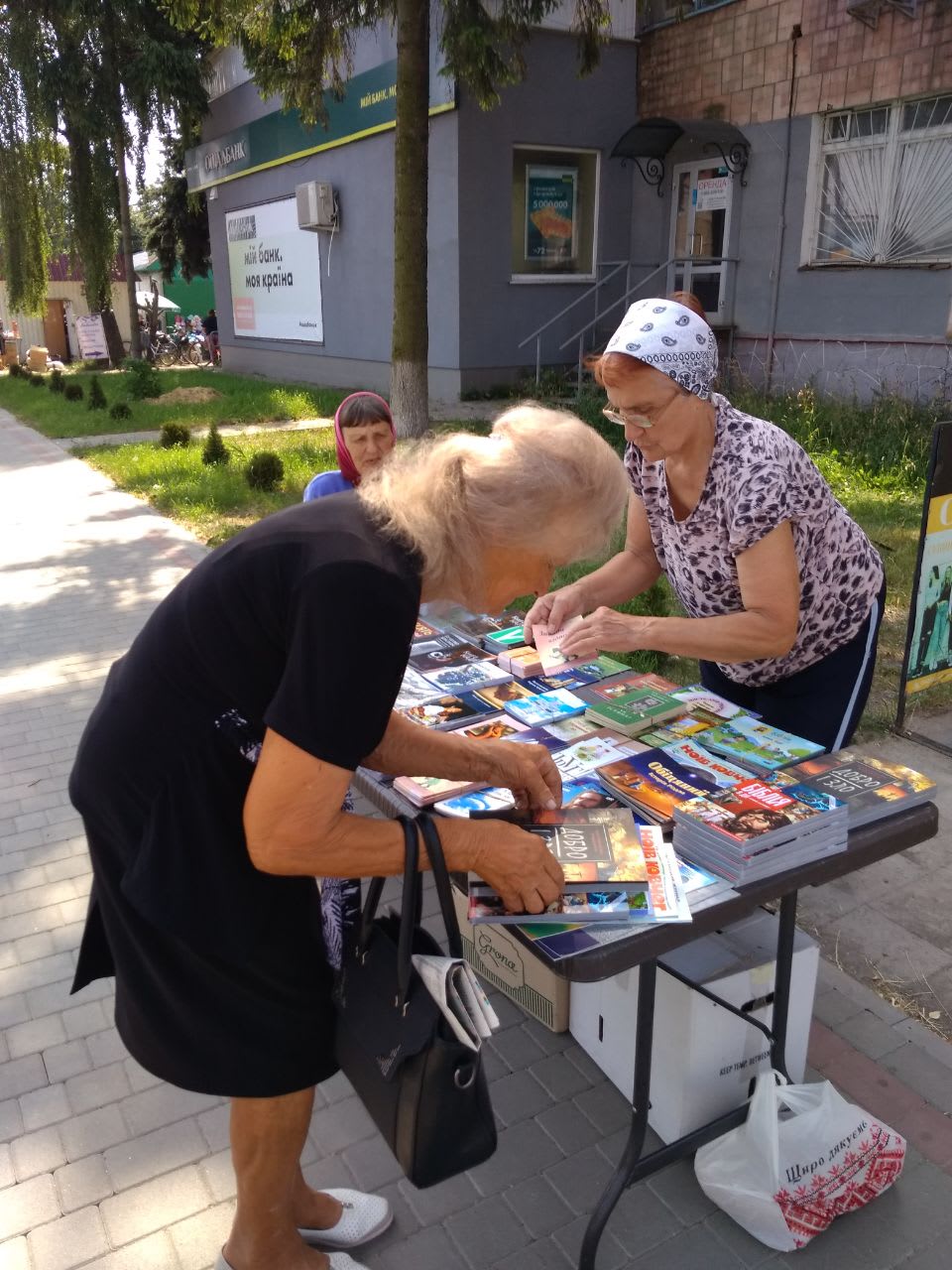 Two older women browse Christian books and literature laid out on an outdoor table on a city sidewalk