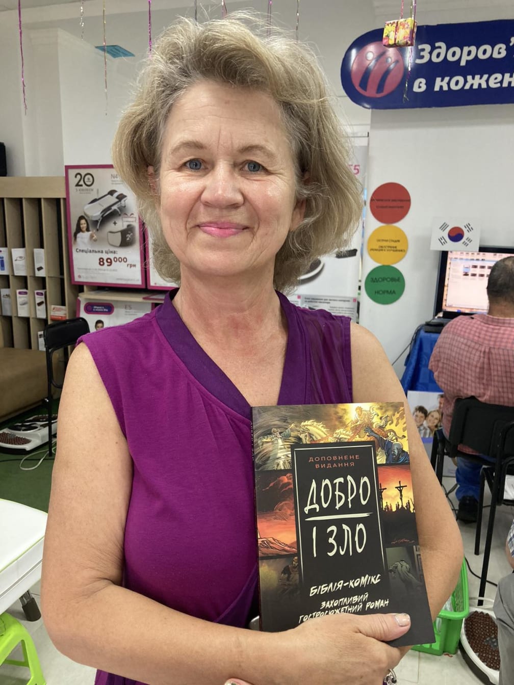 A smiling woman in a store holds a Добро і Зло (Good and Evil) book against her chest